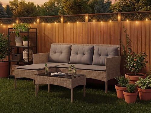 Conjunto lounge de jardim em rattan com almofadas cinzentas, incluindo um sof&aacute; de tr&ecirc;s lugares, um puff e uma mesa de centro num relvado com luzes decorativas e vasos de plantas