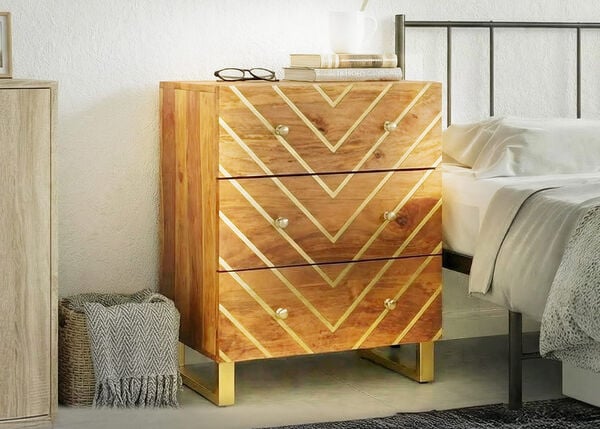 Mesa de cabeceira em madeira com tr&ecirc;s gavetas e padr&atilde;o geom&eacute;trico dourado, colocada ao lado de uma cama num quarto luminoso.