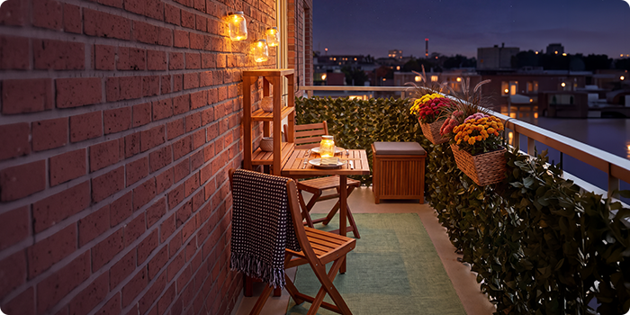 Varanda pequena com cadeiras e mesa de madeira, iluminada por lanternas e velas, decorada com flores em vaso e plantas, com vista para a silhueta da cidade à noite.