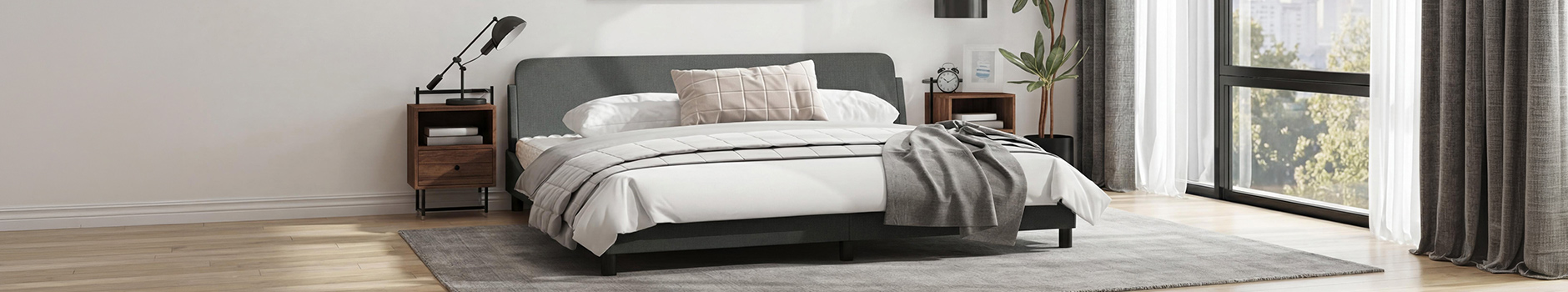 Imagem ampla de um quarto moderno com uma estrutura de cama cinzento-escura, roupa de cama branca e uma almofada bege com padrão em grelha. O quarto tem duas mesas de cabeceira de madeira com candeeiros, uma planta alta em vaso e janelas grandes com cortinados cinzentos e brancos, com vista para árvores verdes.