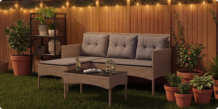  Conjunto lounge de jardim em rattan com almofadas cinzentas, incluindo um sofá de três lugares, um puff e uma mesa de centro num relvado com luzes decorativas e vasos de plantas
