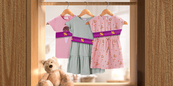 Roupa infantil em cabides envolta numa faixa promocional roxa com ícones de desconto amarelos, pendurada num varão dentro de casa. Clique para ver os produtos em oferta.