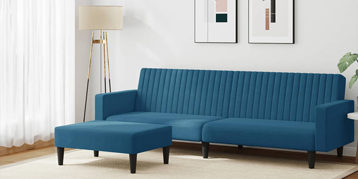 Um sofá modular moderno em veludo azul, com acolchoado vertical em canais e um pufe a condizer. O sofá tem pernas de madeira preta e está numa sala de estar luminosa, com quadros na parede e um candeeiro de pé no canto.